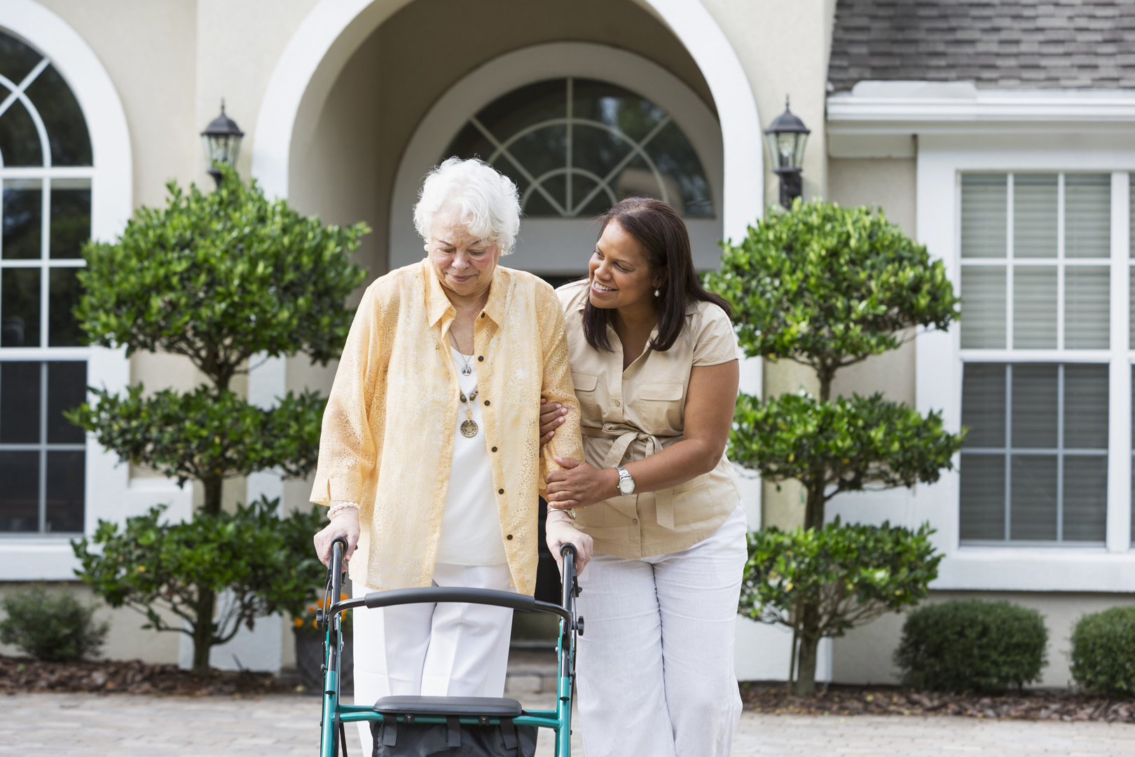 Senior Hispanic woman (70s) with caregiver (50s) in front of house.